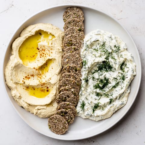 A close-up of vibrant Easy Dips: creamy yogurt beside hummus with herby garnishes and crackers.